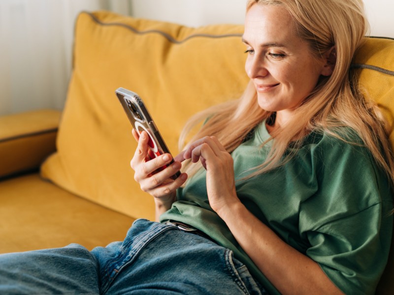 Frau auf dem Sofa, mit Handy in der Hand, die lächelt.