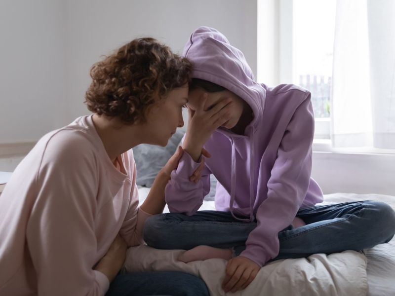 Mutter tröstet ihre Tochter, die weinend auf dem Bett sitzt.