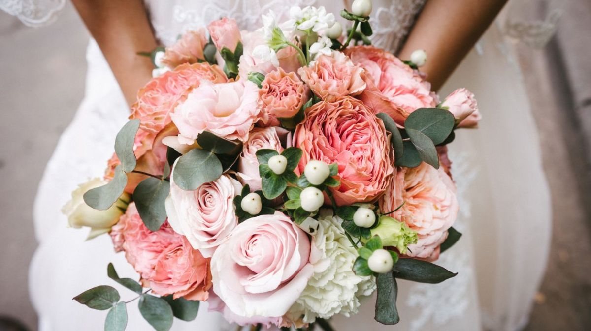 Braut hält einen Brautstrauß mit Rosen und Pfingstrosen vor sich.