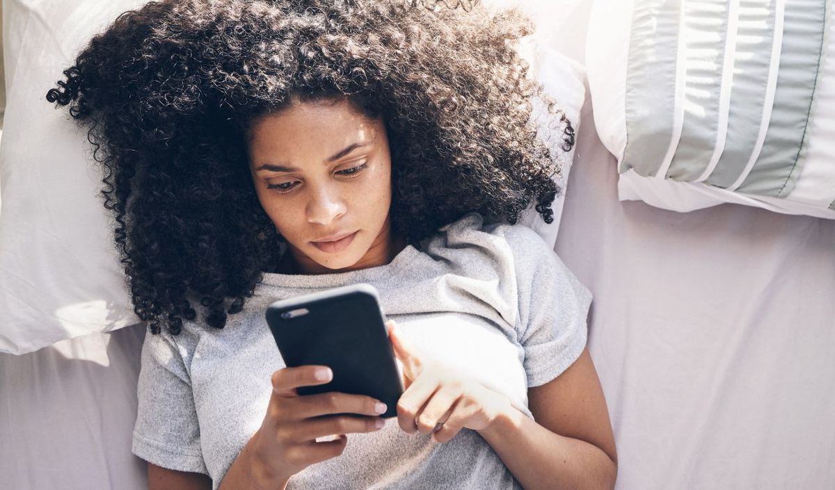 Frau mit dunklen Locken liegt im Bett und schaut auf ihr Handy.