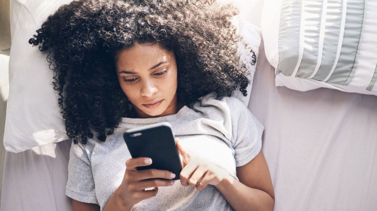 Frau mit dunklen Locken liegt im Bett und schaut auf ihr Handy.