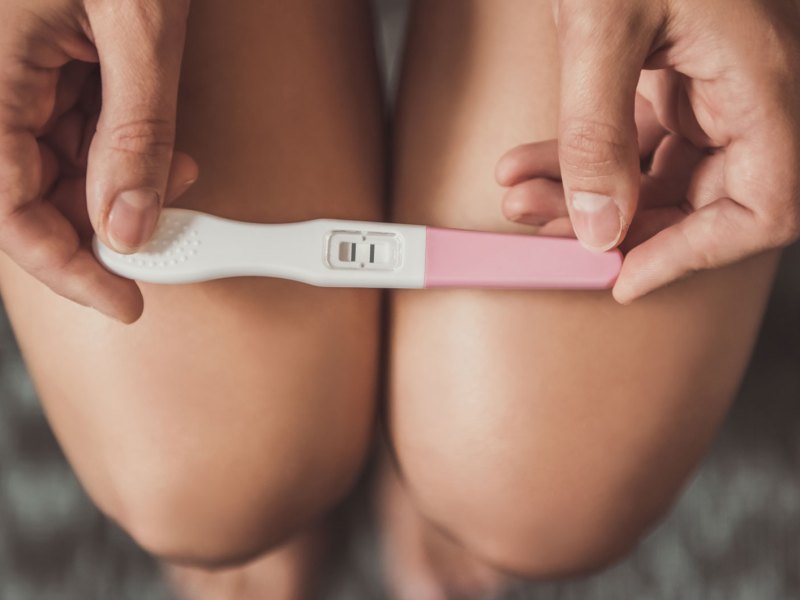 Nahaufnahme eines positiven Schwangerschaftstests, gehalten von zwei Frauenhänden, die auf den nackten Oberschenkeln liegen.