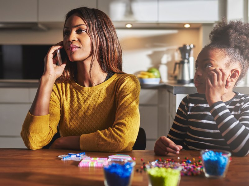 Mutter und Tochter sitzen am Küchentisch. Die Mutter telefoniert mit ihrem Smartphone und die Tochter sitzt daneben und wartet darauf, dass die Mutter wieder Zeit für sie hat.