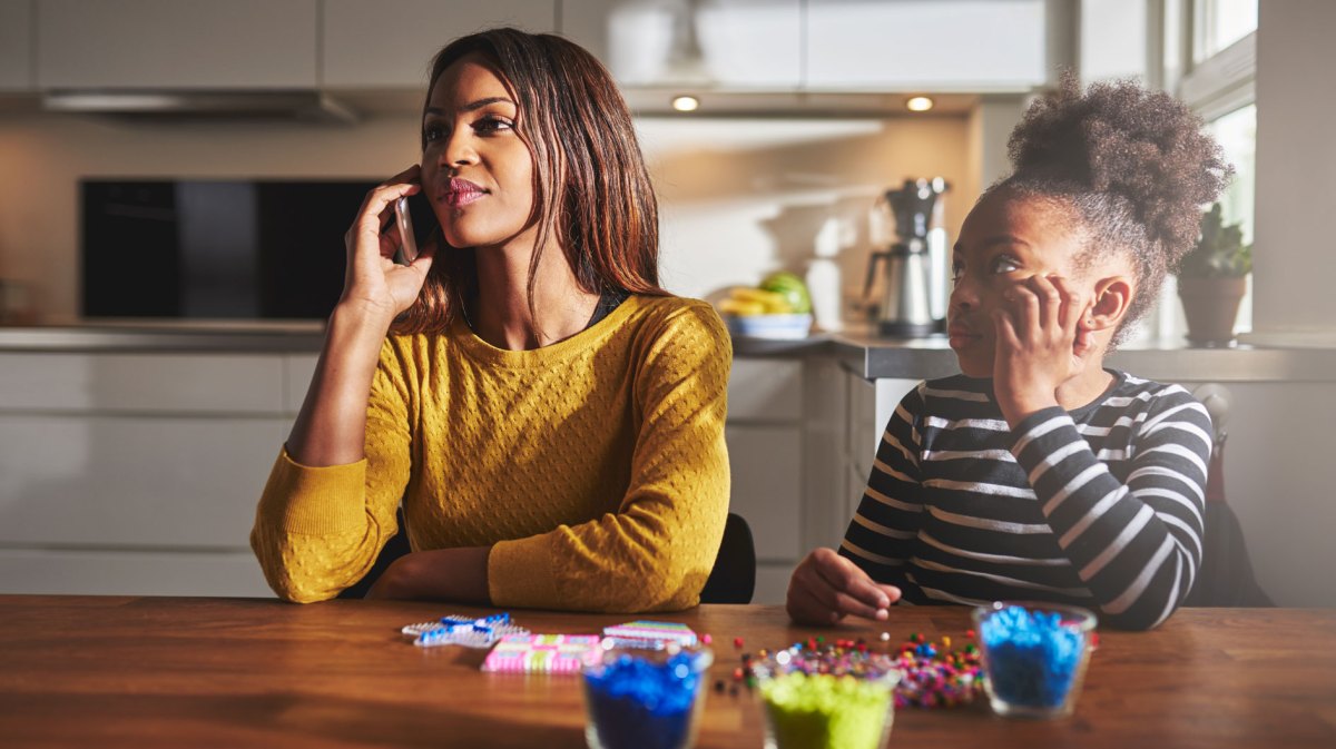 Mutter und Tochter sitzen am Küchentisch. Die Mutter telefoniert mit ihrem Smartphone und die Tochter sitzt daneben und wartet darauf, dass die Mutter wieder Zeit für sie hat.