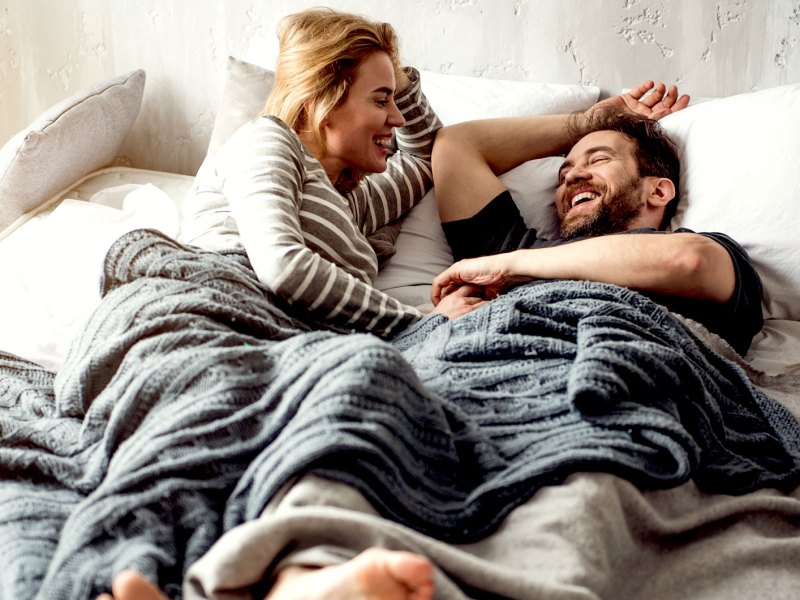 Mann und Frau liegen entspannt und lachend im Bett nebeneinander und unterhalten sich.