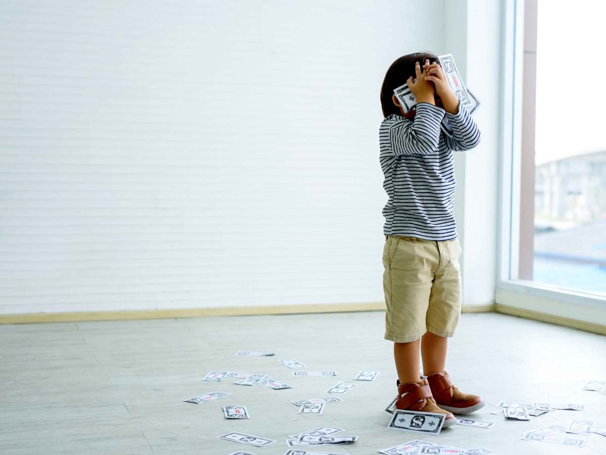 Kleiner Junge steht in einem leeren Zimmer und hält sich seine Hände, die voll mit Geldscheinen sind, vor das Gesicht. Um ihn herum liegen weitere Scheine auf dem Boden.