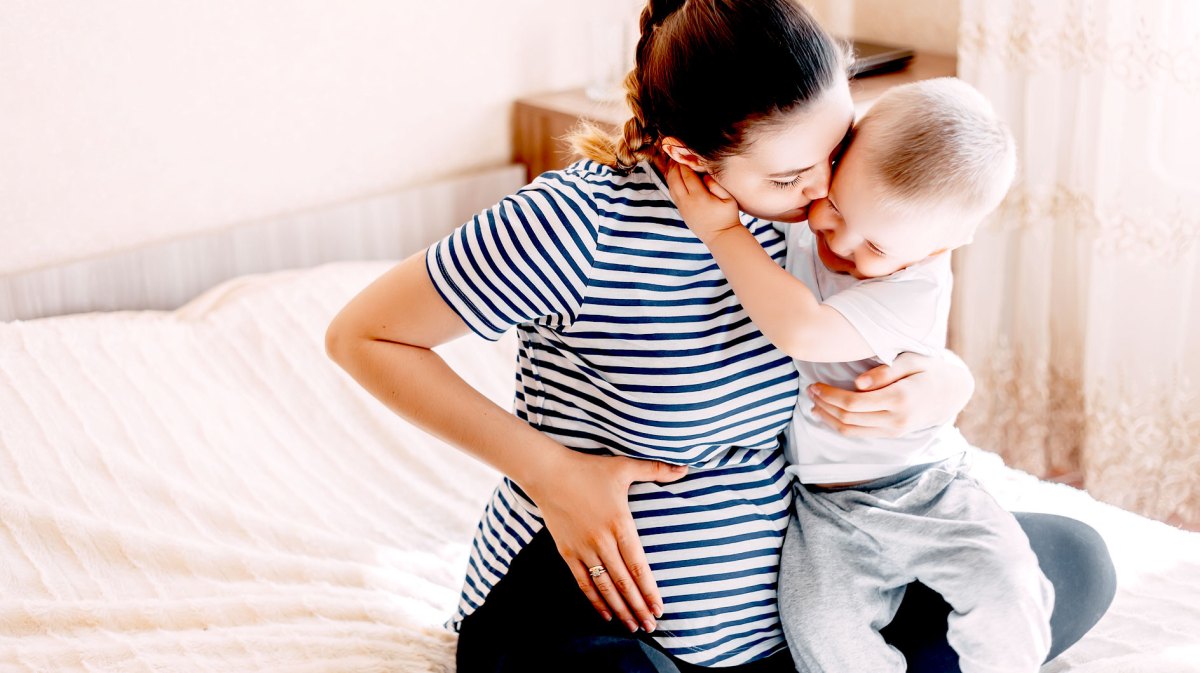 Hochschwangere Mutter und Kleinkind sitzen auf einem großen Bett und kuscheln.