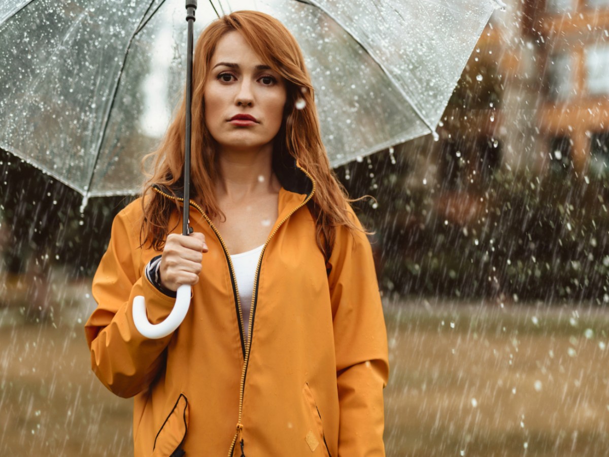 Frau in orangem Regenmantel, steht mit Schirm im Regen.