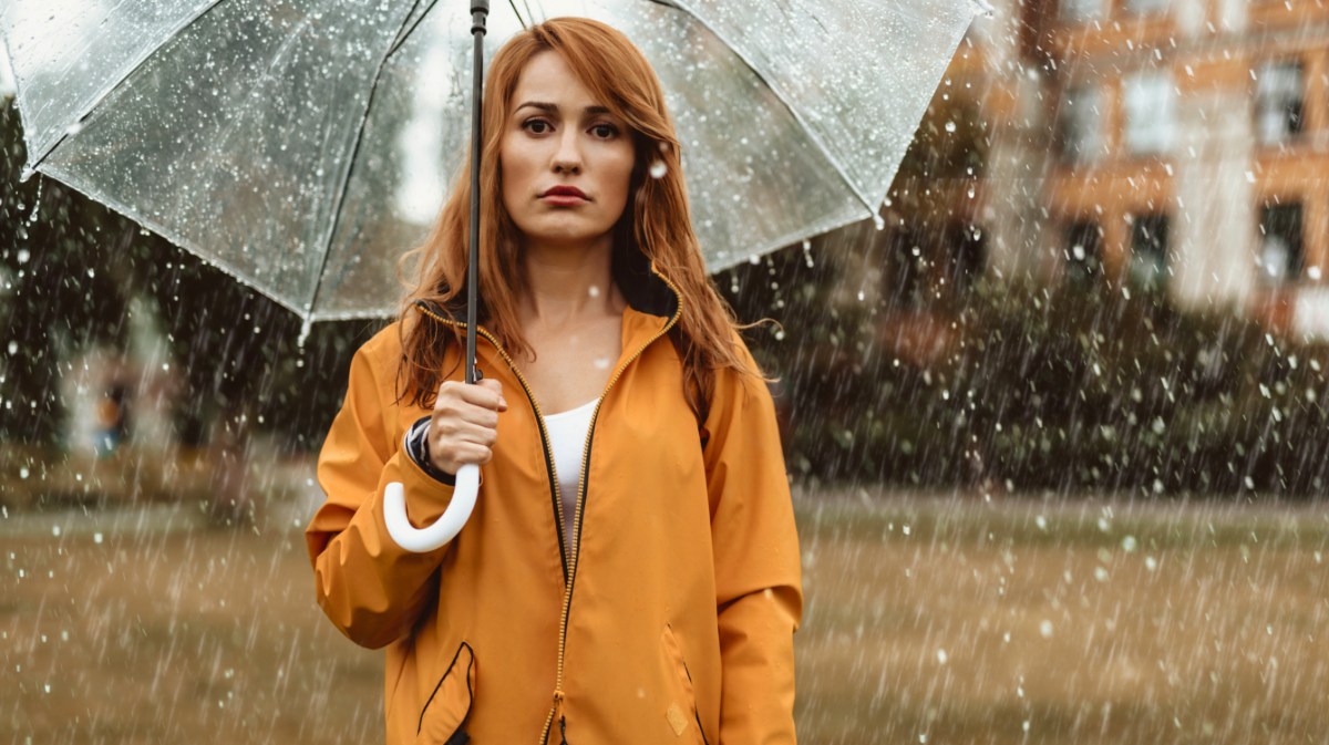 Frau in orangem Regenmantel, steht mit Schirm im Regen.