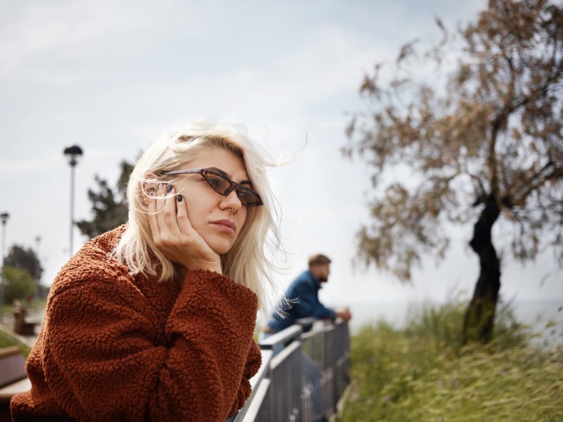 Frau schaut mit Sonnenbrille gelangweilt von einer Brücke hinunter, im Hintergrund ein Mann der ebenfalls herunterschaut