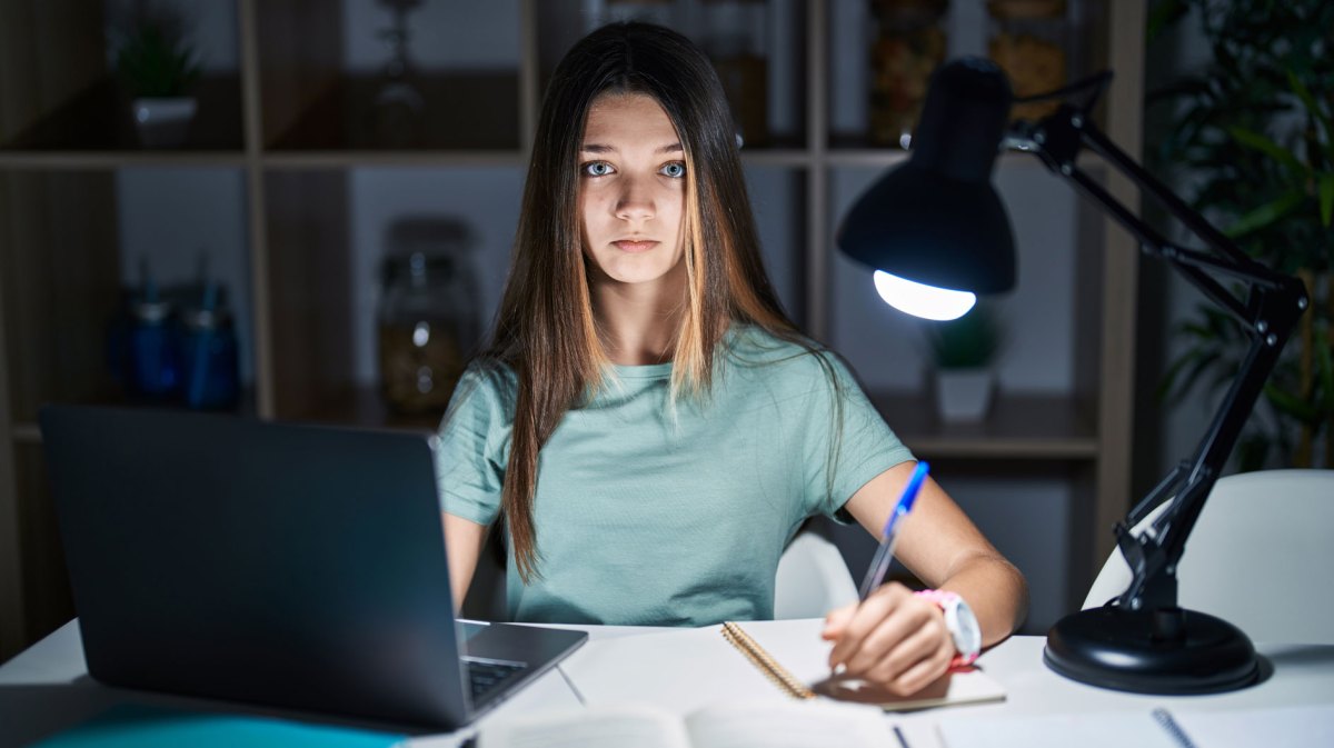 Teenagermädchen sitzt im Dunkeln an ihrem Schreibtisch, der von einer Schreibtischlampe beleuchtet wird und lernt.