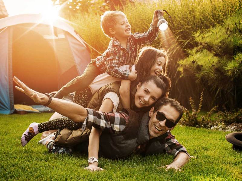 Vierköpfige Familie liegt übereinander gestapelt auf dem Gras und albert herum, hinter sich ein Zelt. Der Jüngste ganz oben hält eine Taschenlampe.