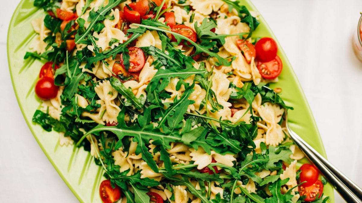 Nudelsalat mit Farfalle, Rucola und Tomaten in einer grünen Servierschüssel.