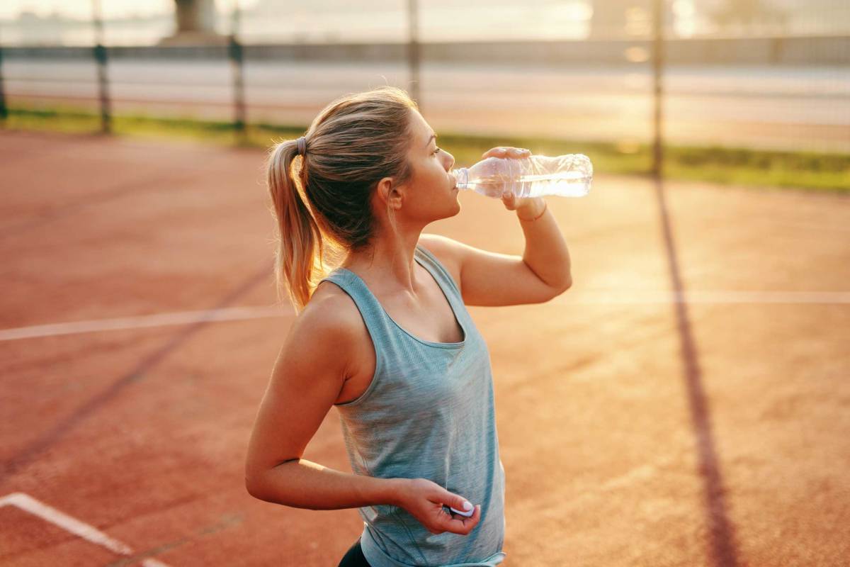 Sportliche blonde Frau steht auf dem Platz und trinkt Wasser nach dem Morgensport.