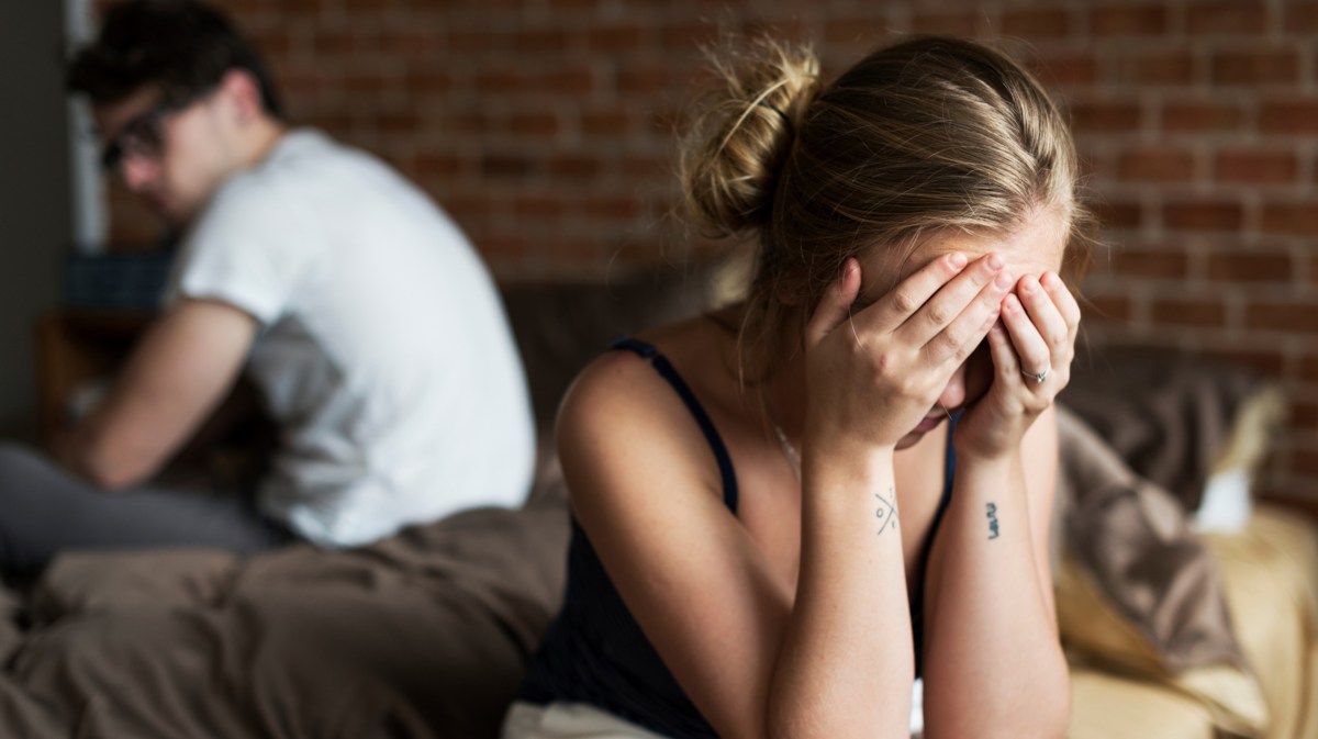 Frau sitzt mit Händen vor dem Gesicht auf Bettkante mit verschwommenen Bild eines Mannes im Hintergrund