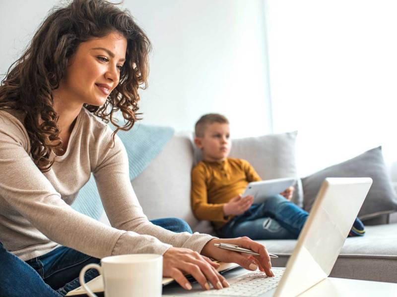 Mutter sitzt im Wohnzimmer am Laptop, neben sich eine Tasse Kaffe. Im Hintergrund sieht man den Sohn an einem Tablet auf der Couch sitzen.