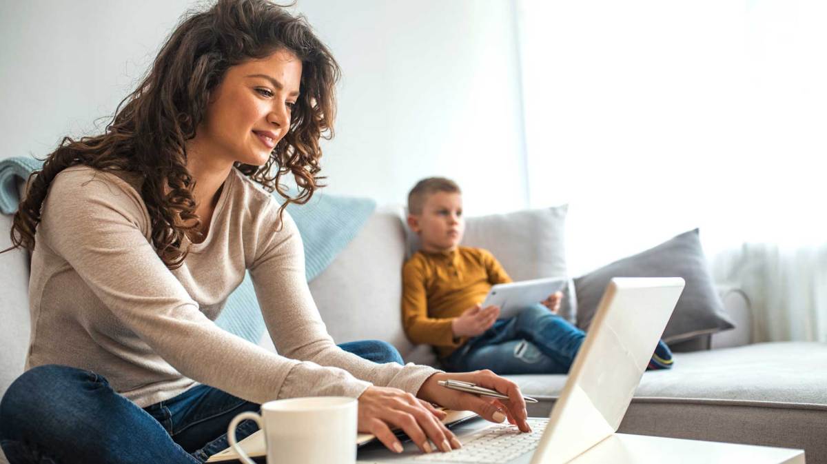 Mutter sitzt im Wohnzimmer am Laptop, neben sich eine Tasse Kaffe. Im Hintergrund sieht man den Sohn an einem Tablet auf der Couch sitzen.