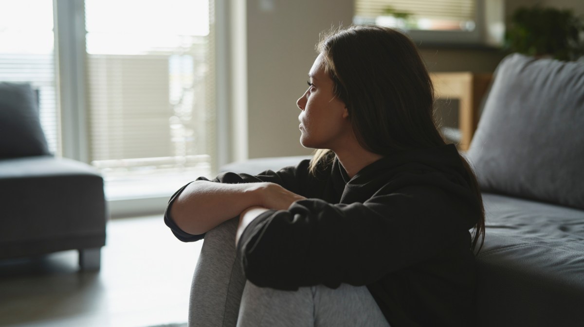 Frau schaut nachdenkllich und traurig aus Fenster, während sie auf Holzboden vor Couch sitzt