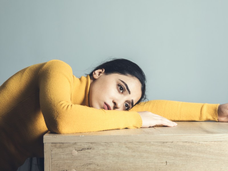 Frau in gelbem Pullover, die auf einem Tisch mit ausgebreiteten Armen liegt und bedrückt nach vorne schaut und in Nostalgie schwirrt