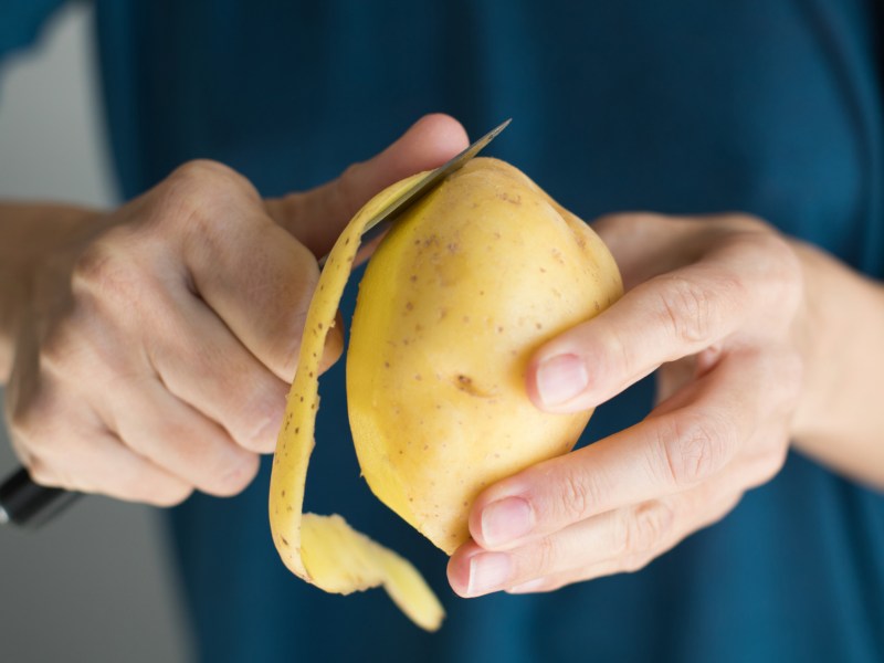 Person mit blauem Oberteil hält Kartoffel in den Händen und schält sie.