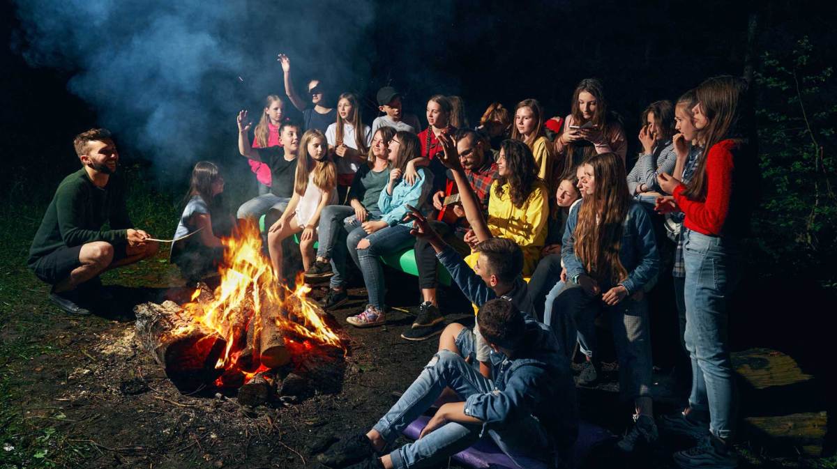 Zahlreiche Kinder sitzen und stehen um ein Lagerfeuer herum. Vor dem Feuer hockt auch ein Erwachsener.