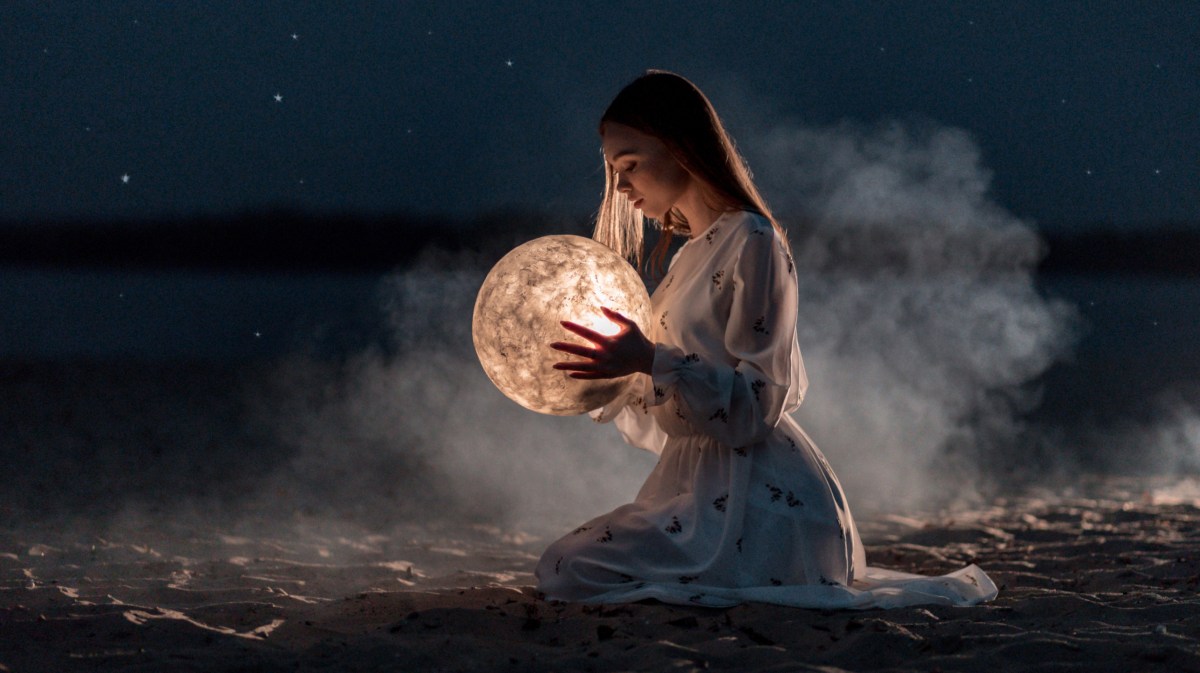 Frau sitzt an Strand und hält Mond in der Hand.