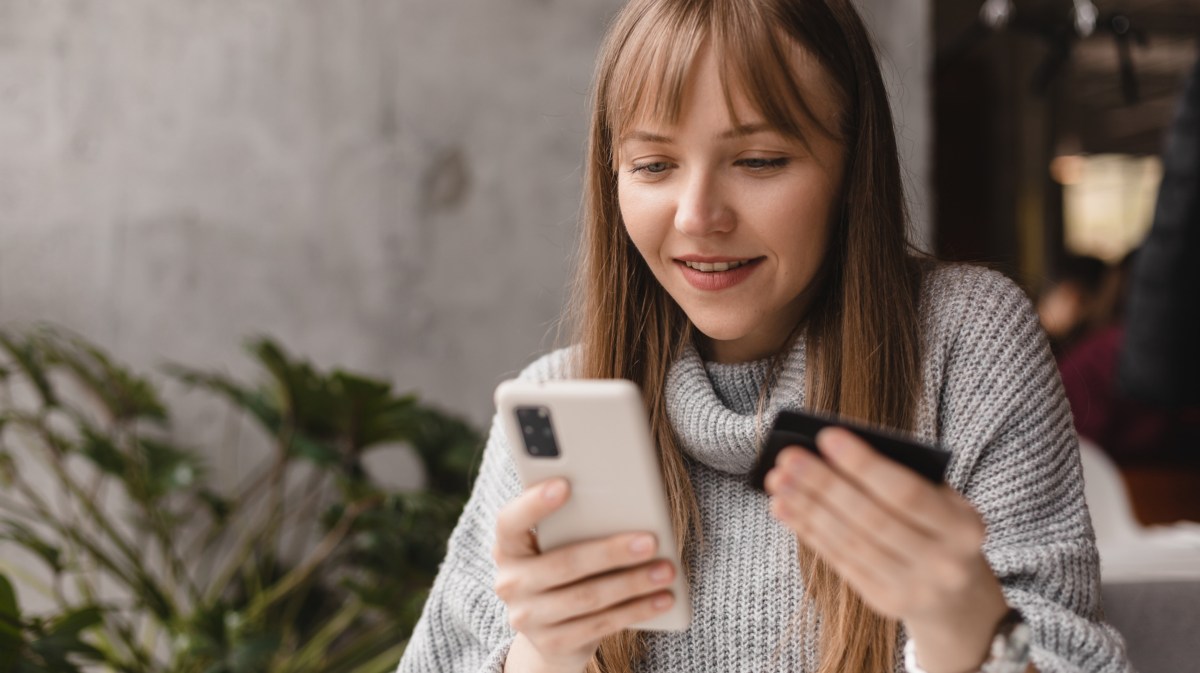 Junge Frau hält eine Kreditkarte in der Hand und ihr Handy in der anderen Hand und schaut gespannt auf das Telefon