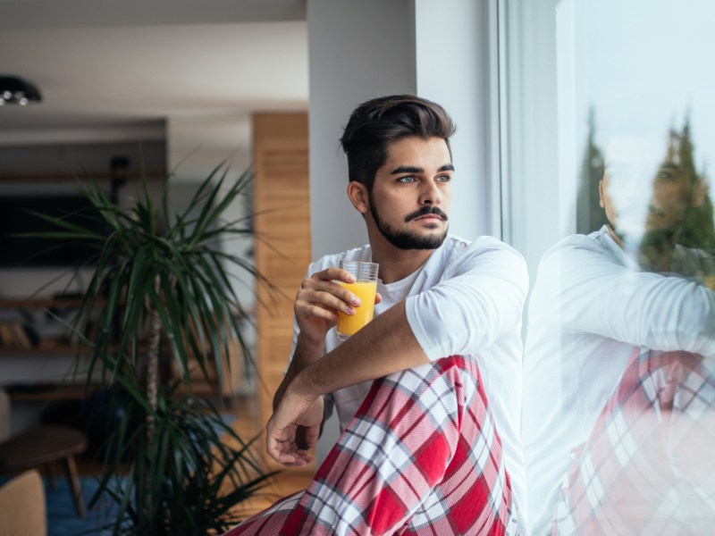 Mann, der mit Schlafanzug und Kaffee-Tasse in der Hand am Fenster sitzt und nachdenklich herausschaut