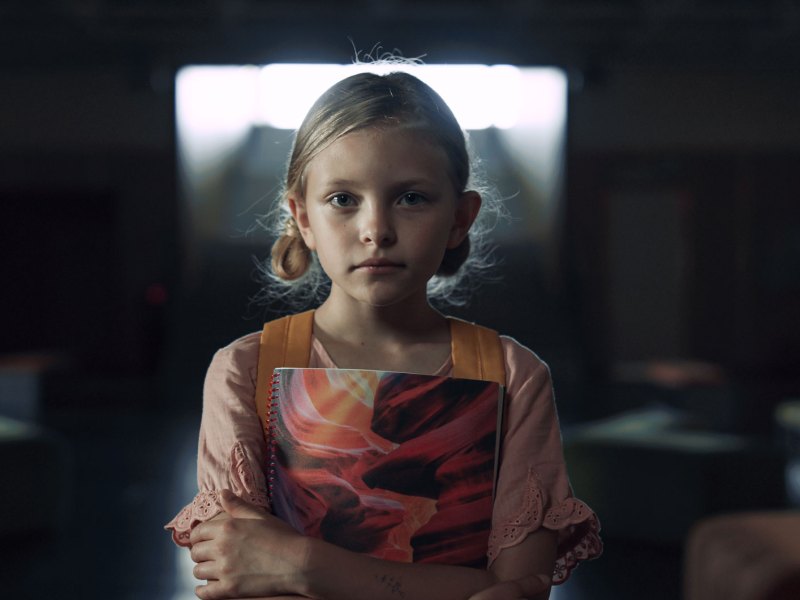 Mädchen, ca. 10 Jahre, steht mit Rucksack und Schreibblock in einem verdunkelten Klassenzimmer und schaut neutral, aber doch besorgt in die Kamera.