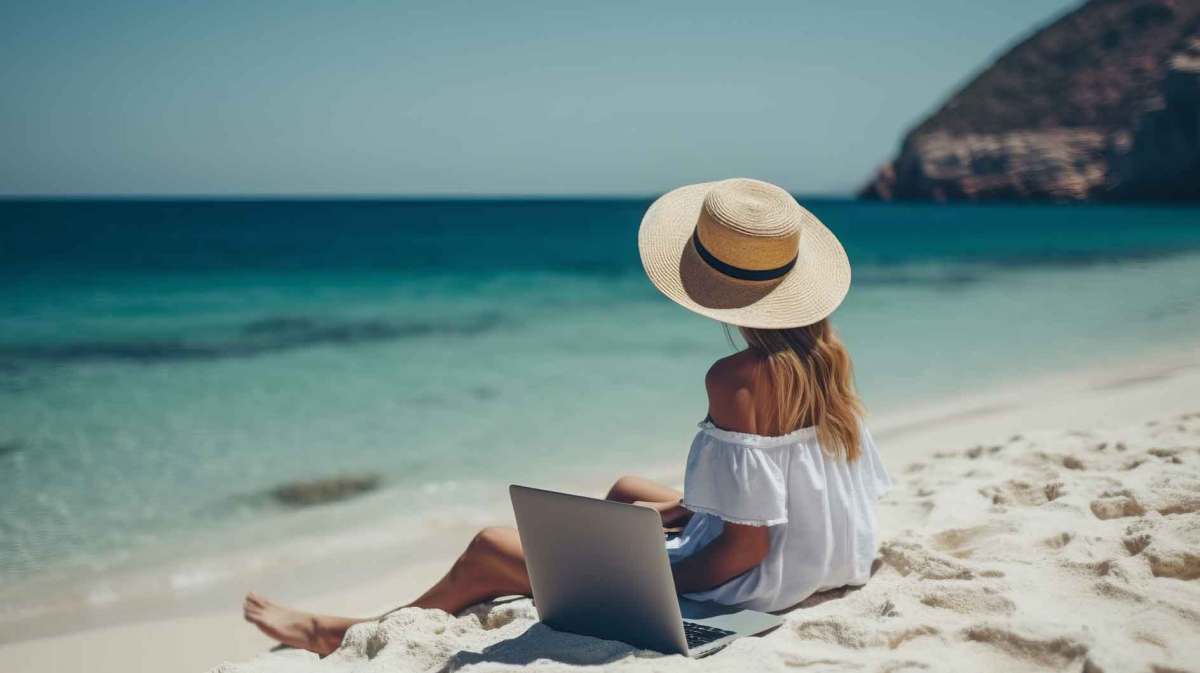 Junge Frau mit Sonnenhut sitzt am Strand mit ihrem Laptop und hat Urlaub.