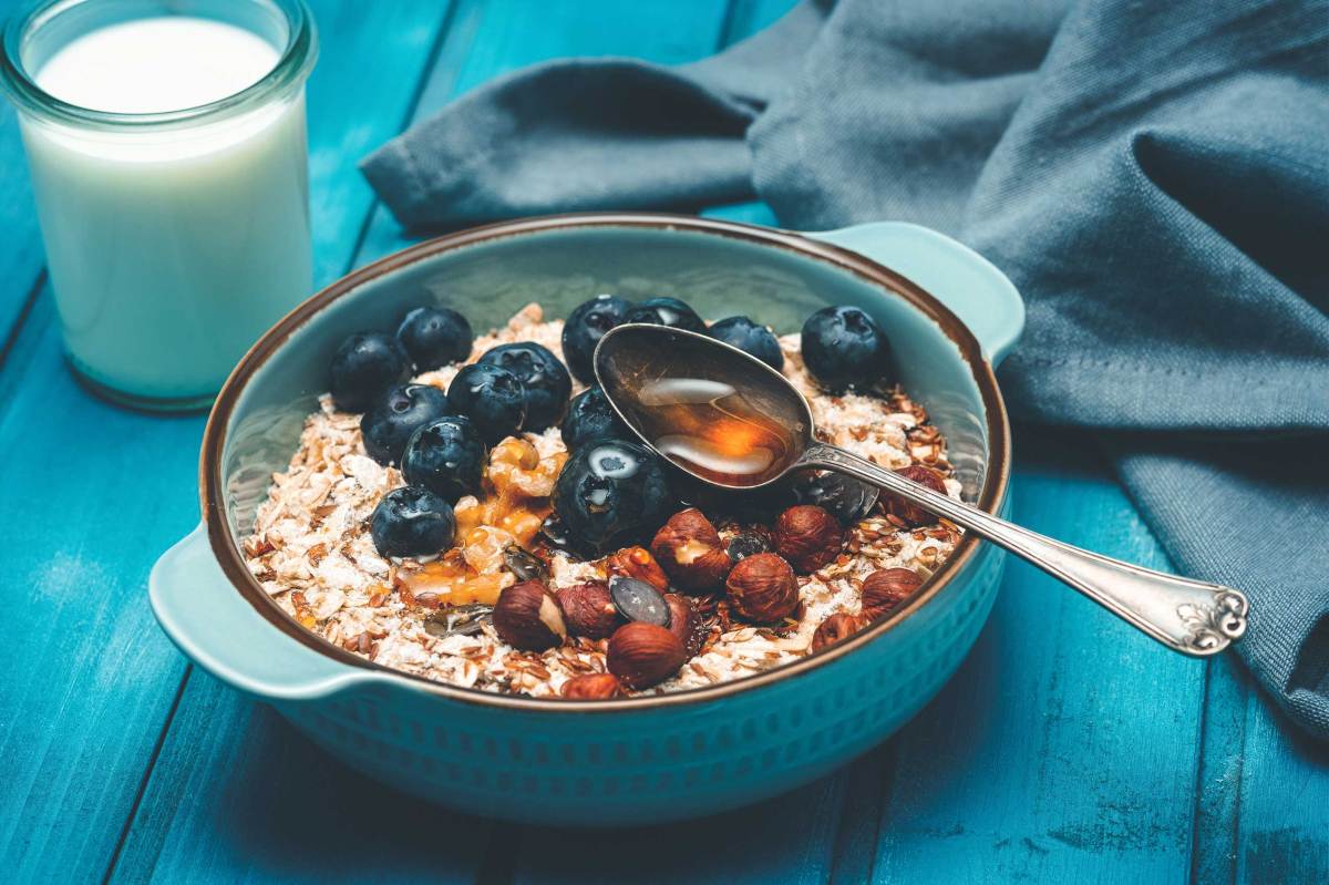 Blaubeeren und Nüsse auf Haferflocken in einer türkisfarbenen Schale, daneben ein Glas Milch.