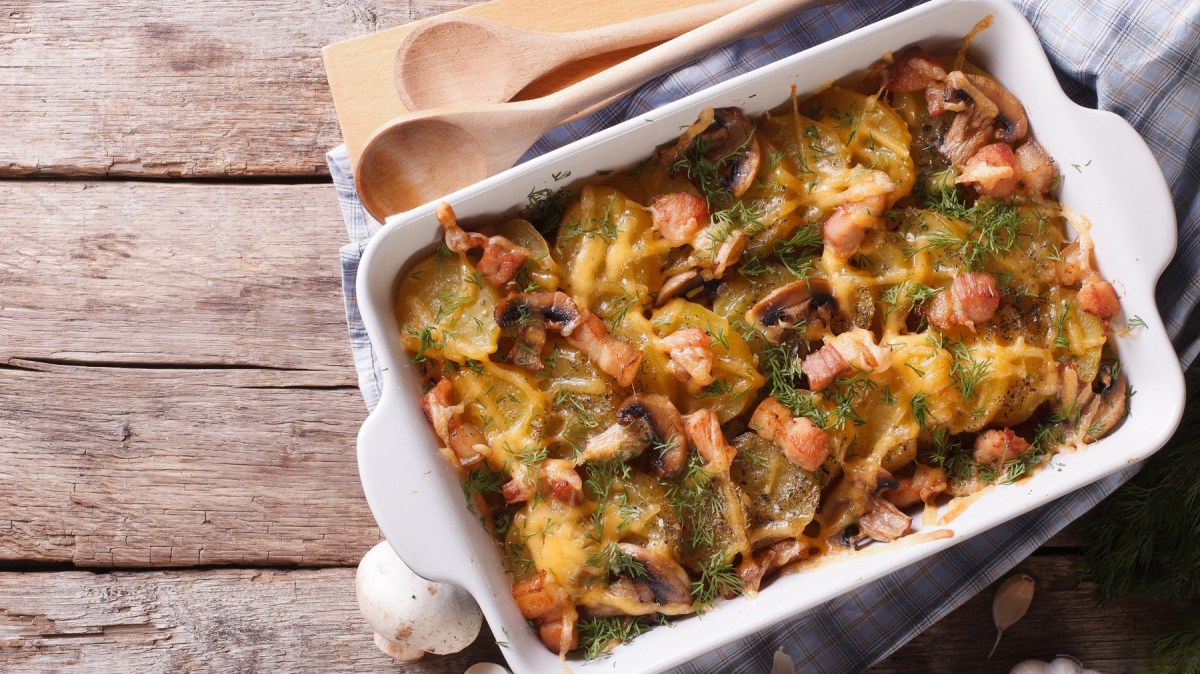 Eine weiße Auflaufform auf einem hellen Holztisch mit dem Gratin und einem Bund Kräuter sowie Pilzen und Knoblauchknollen.