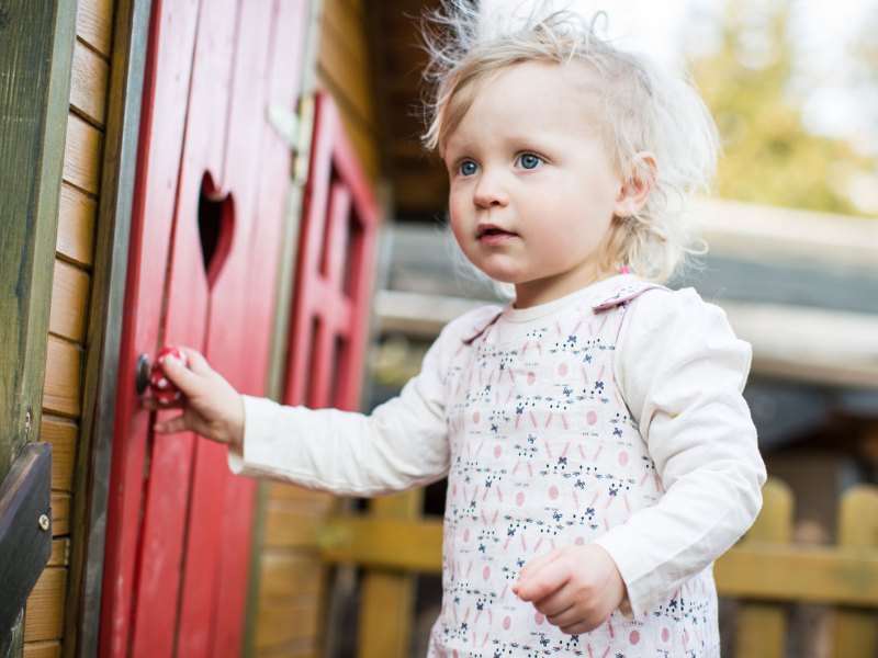 Kleines Mädchen, ca. 2 Jahre, steht draußen an einem Spielhaus mit roter Tür und einem herzförmigen Guckloch.