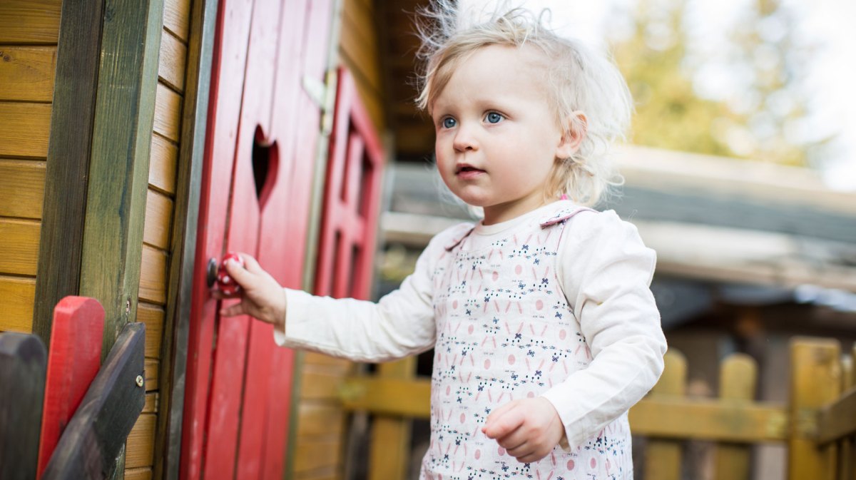 Kleines Mädchen, ca. 2 Jahre, steht draußen an einem Spielhaus mit roter Tür und einem herzförmigen Guckloch.