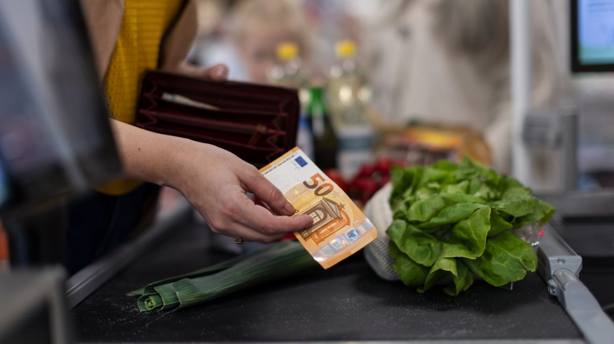 Frau, die einen 50-Euro-Schein in der Hand hält und auf dem Kassenband eine Gurke und einen Salatkopf gelegt hat