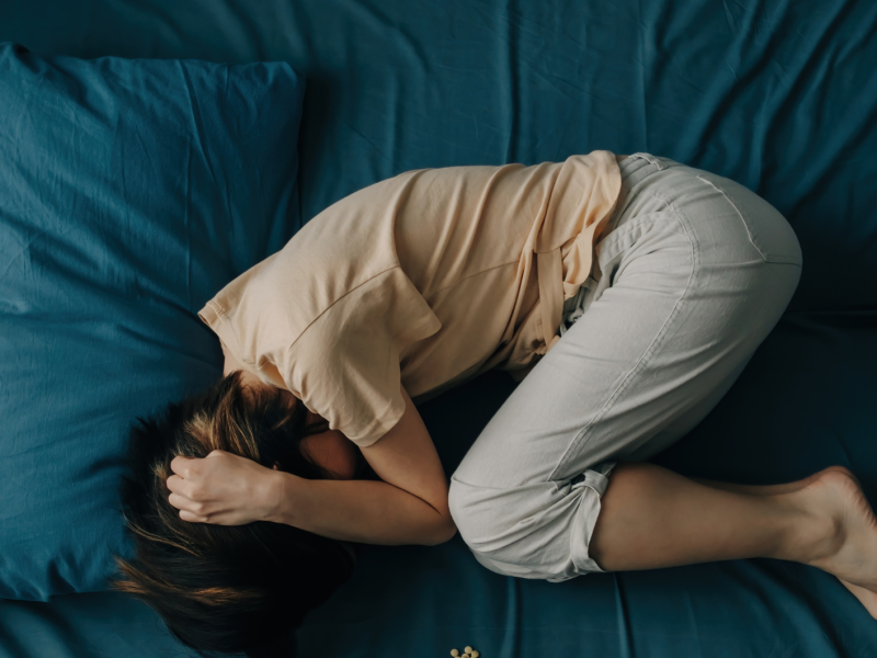 Eine junge Frau mit dunklem Haar liegt zusammengerollt in Seitenlage auf einem blau bezogenen Bett und hält ihren Kopf mit der Hand.