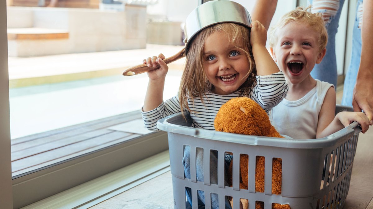 Bruder und Schwester sitzen in einem Wäschekorb und werden durch einen Flur geschoben. Das Mädchen trägt einen Topf auf dem Kopf.