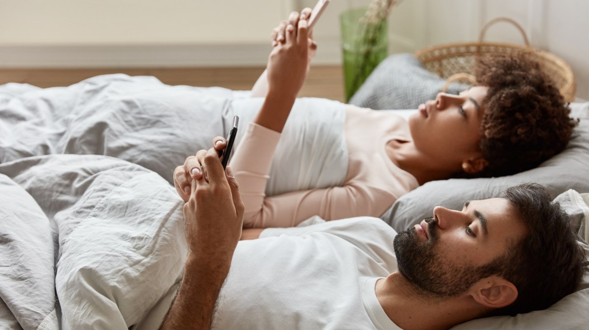 Mann und Frau die gemeinsam nebeneinander im Bett liegen und gelangweilt auf ihr Handy schauen