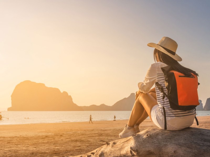 Frau mit Rucksack und Hut sitzt auf Stein am Strand und schaut den Sonnenuntergang