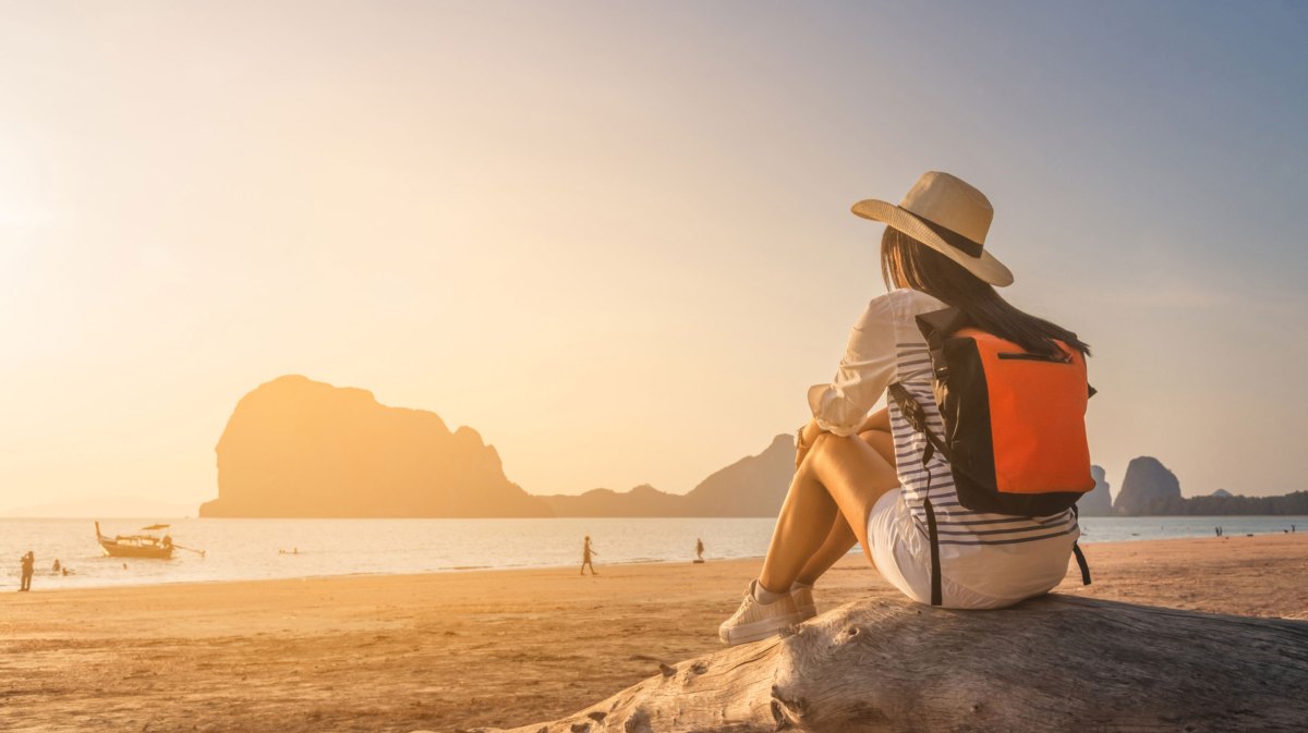 Frau mit Rucksack und Hut sitzt auf Stein am Strand und schaut den Sonnenuntergang