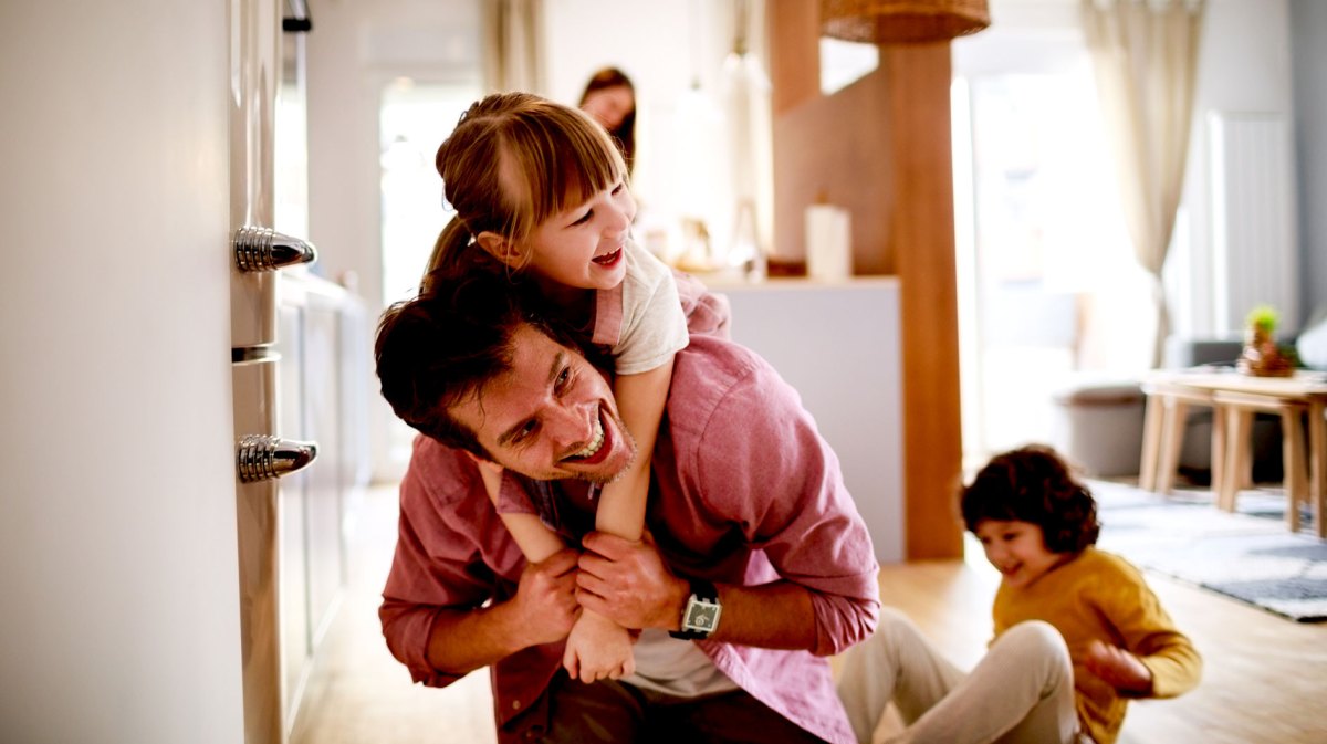 Vater tobt mit seinen beiden Kindern im Haus. Die Tochter trägt er gerade auf den Schultern, der Sohn sitzt im Hintergrund lachend auf dem Boden.