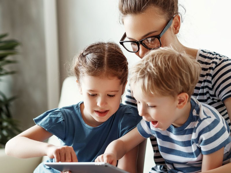 Frau und zwei Kinder sitzen gemeinsam auf dem Sofa und schauen etwas auf dem Tablet.