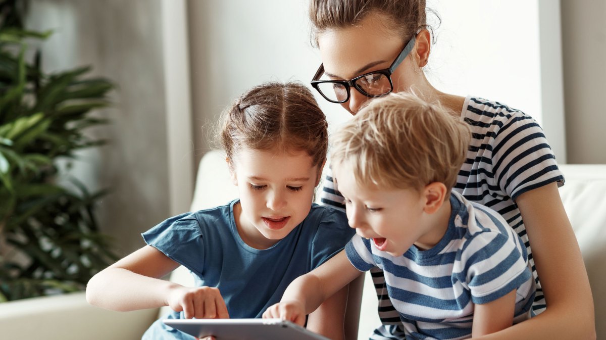 Frau und zwei Kinder sitzen gemeinsam auf dem Sofa und schauen etwas auf dem Tablet.