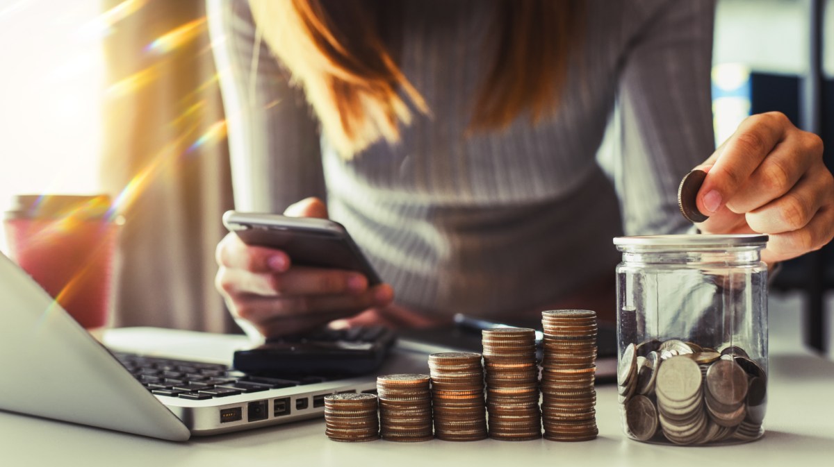 Frau schaut auf Handy und hat mehrere Stapel Münzgeld vor sich auf Tisch liegen.