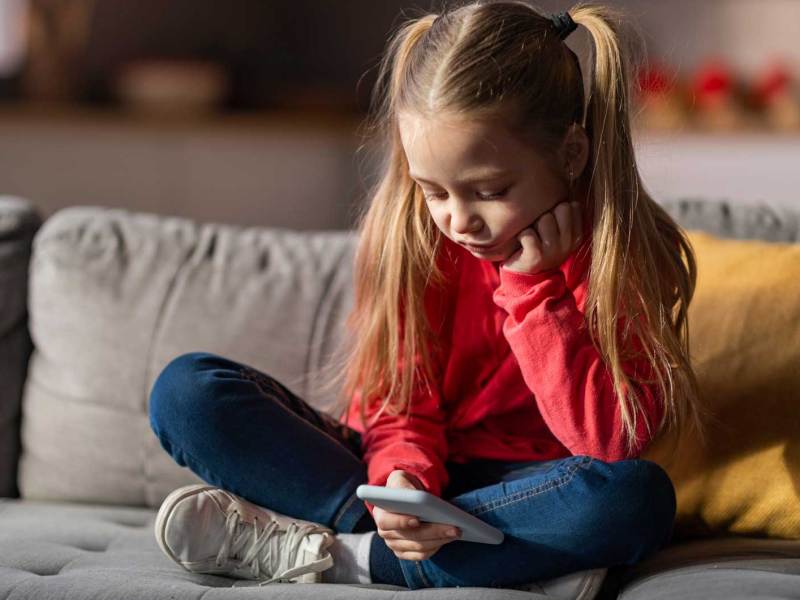 Mädchen, ca. 6 Jahre alt, sitzt im Schneidesitz auf dem Sofa und spielt an einem Handy.