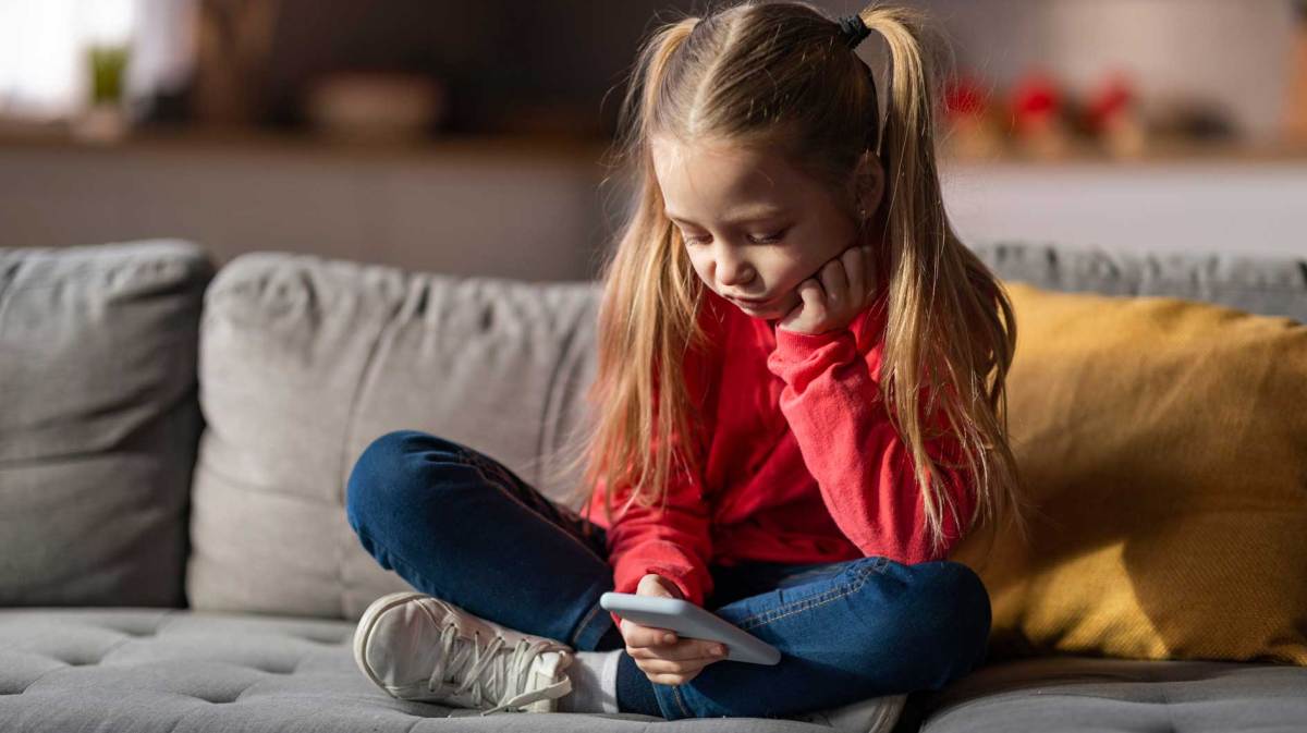 Mädchen, ca. 6 Jahre alt, sitzt im Schneidesitz auf dem Sofa und spielt an einem Handy.