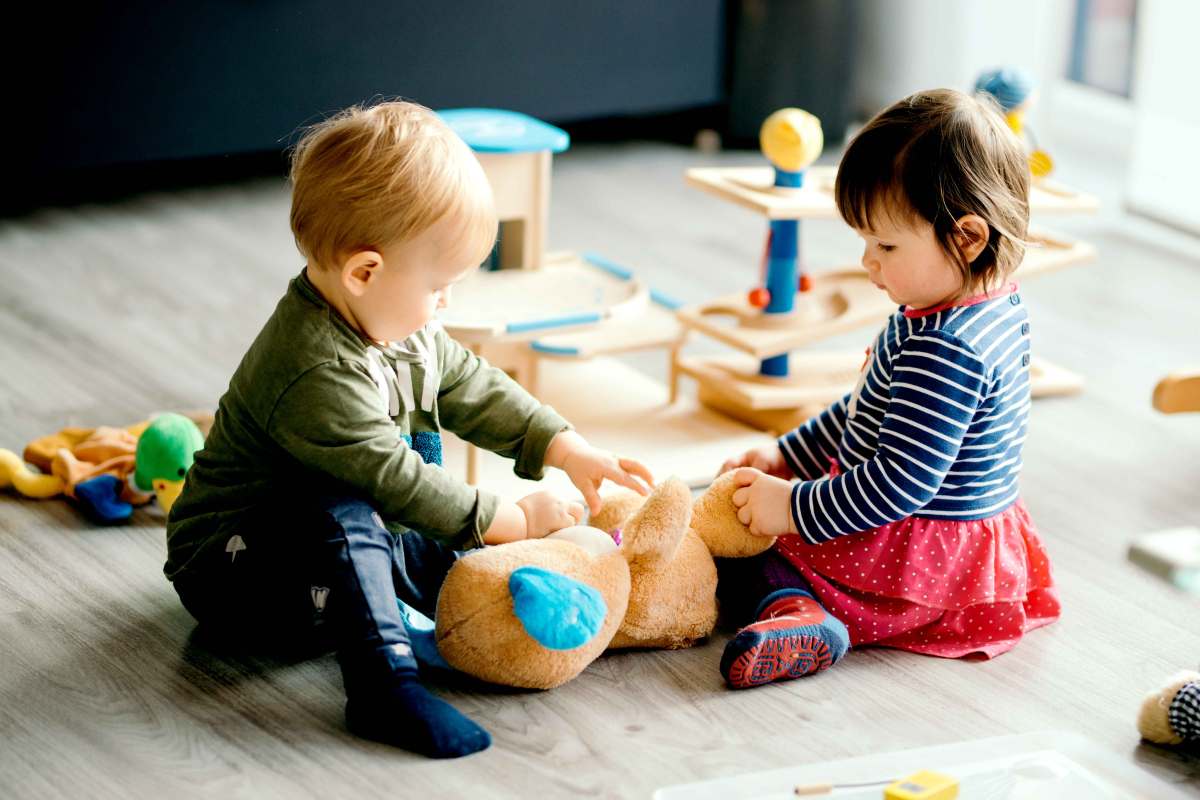 Zwei Kleinkinder sitzen auf dem Fußboden und spielen mit einem großen Plüschteddy.