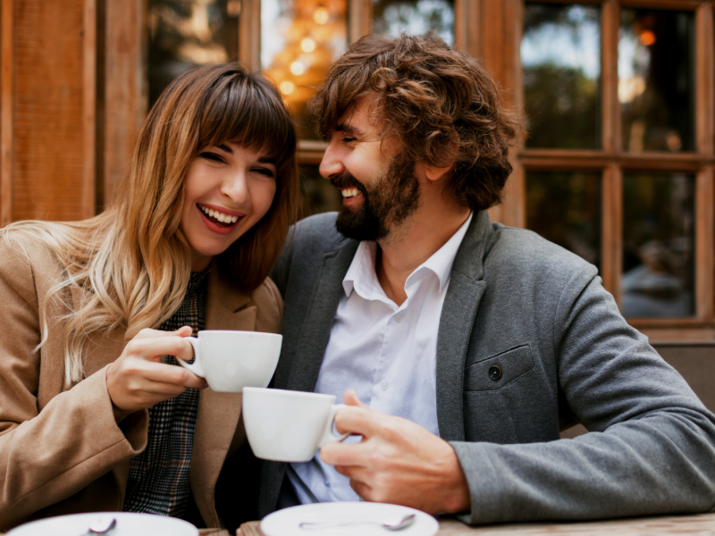 Eine junge Frau mit braunen Haaren und ein bärtiger Mann lachen sich an, während sie draußen vor einem Café Kaffee trinken.