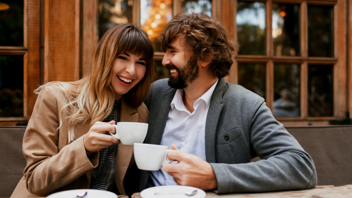 Eine junge Frau mit braunen Haaren und ein bärtiger Mann lachen sich an, während sie draußen vor einem Café Kaffee trinken.