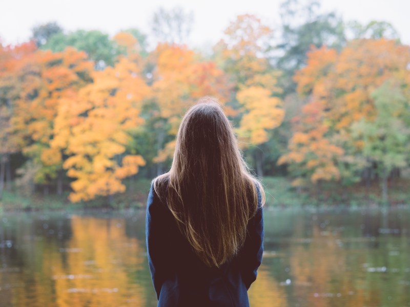 Junge Frau mit braunen Haaren steht vor einem See im Herbst und hat der Kamera den Rücken zugwandt.